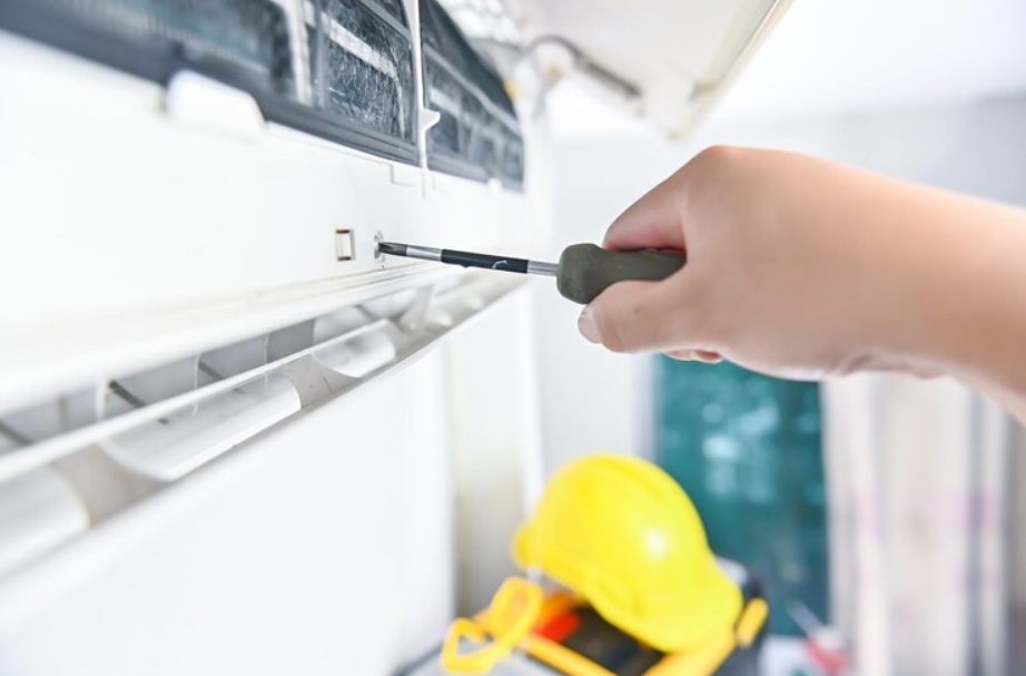 Hand using screwdriver to service mini split air conditioning unit with yellow hard hat and tools in background, relevant to HVAC maintenance and troubleshooting.